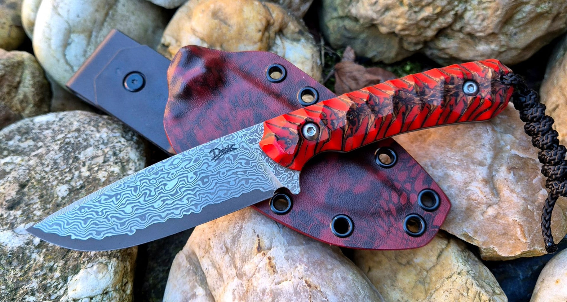 Damaškový nůž položený na Kydex pouzdru, zdůrazňující texturu čepele a kontrastní červenou rukojeť – prémiový custom nůž. custom nůž, EDC nůž, damašek, lovecký nůž, Kydex pouzdro
